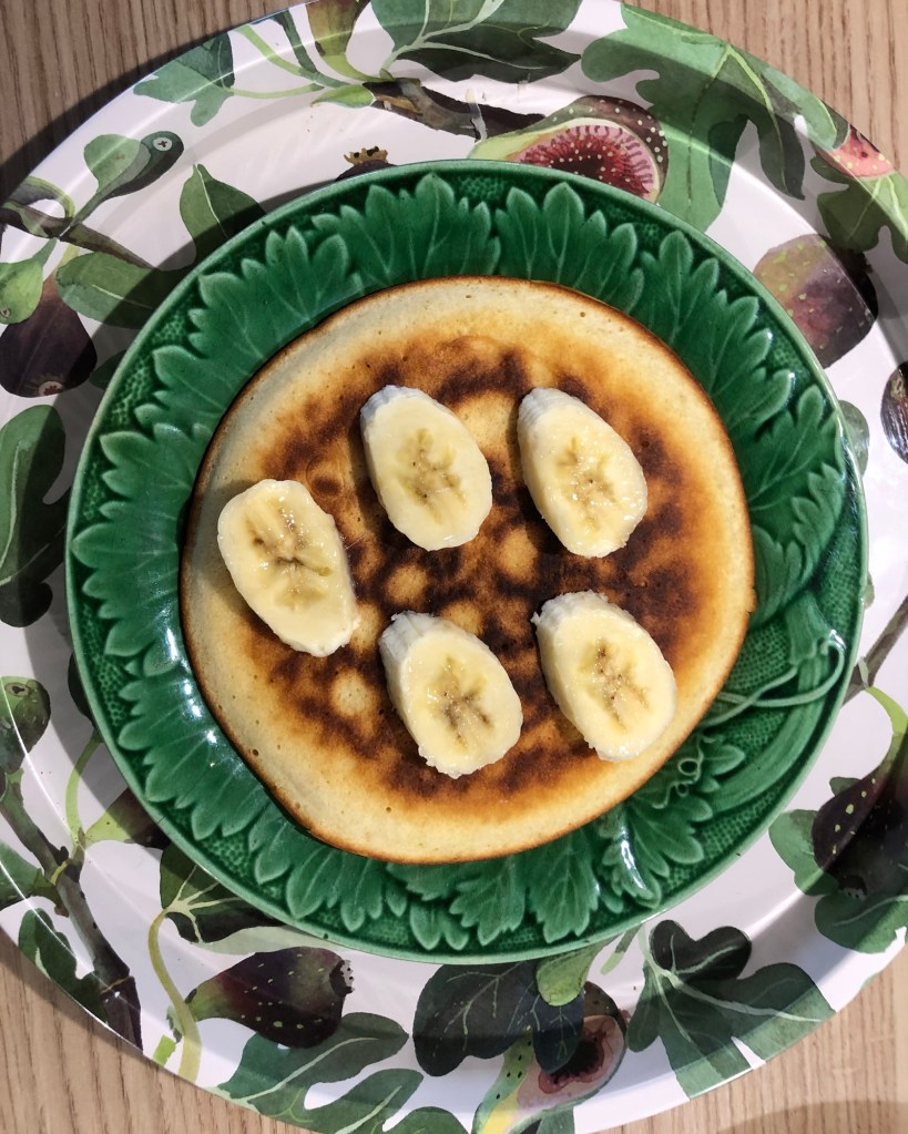 A fluffy American pancake with bananas sliced on top, presented on a green plate and tray, both featuring fig leaf motifs 